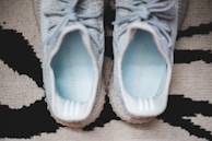 A pair of light grey sneakers with white soles, viewed from above, positioned on a patterned rug. The inside of the sneakers displays light blue insoles featuring text.