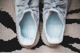 A pair of light grey sneakers with white soles, viewed from above, positioned on a patterned rug. The inside of the sneakers displays light blue insoles featuring text.