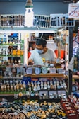 A man wearing a mask works at a stall filled with a vast display of watches and clocks. The stall is brightly lit by a bulb and showcases an array of wristwatches and small clocks, neatly arranged in multiple layers. The man appears to be adjusting or repairing one of the watches.