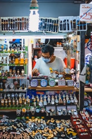 A man wearing a mask works at a stall filled with a vast display of watches and clocks. The stall is brightly lit by a bulb and showcases an array of wristwatches and small clocks, neatly arranged in multiple layers. The man appears to be adjusting or repairing one of the watches.