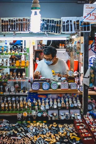 A man wearing a mask works at a stall filled with a vast display of watches and clocks. The stall is brightly lit by a bulb and showcases an array of wristwatches and small clocks, neatly arranged in multiple layers. The man appears to be adjusting or repairing one of the watches.