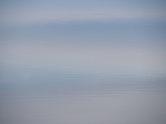 A serene body of water with gentle ripples extending to the horizon. The water reflects a light blue sky with soft gradients, creating a calming and tranquil effect.