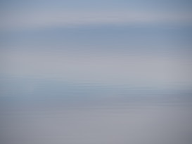 A serene body of water with gentle ripples extending to the horizon. The water reflects a light blue sky with soft gradients, creating a calming and tranquil effect.