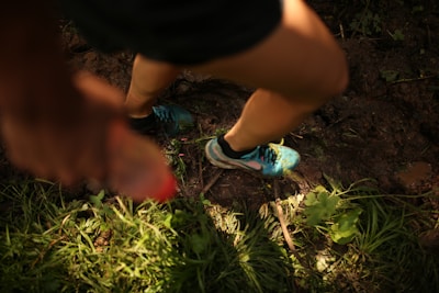 A person is running on a muddy trail surrounded by grass. The focus is on their legs and feet, which are wearing blue running shoes. In the foreground, a hand holding a red-capped bottle is blurred, indicating motion.