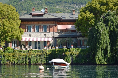 A picturesque lakeside villa surrounded by lush greenery and trees, with a red and white striped awning. The villa has a brick and stone facade, large windows, and outdoor seating. In front, a small boat is moored on calm water, reflecting the serene environment.