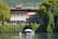 A picturesque lakeside villa surrounded by lush greenery and trees, with a red and white striped awning. The villa has a brick and stone facade, large windows, and outdoor seating. In front, a small boat is moored on calm water, reflecting the serene environment.