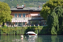 A picturesque lakeside villa surrounded by lush greenery and trees, with a red and white striped awning. The villa has a brick and stone facade, large windows, and outdoor seating. In front, a small boat is moored on calm water, reflecting the serene environment.