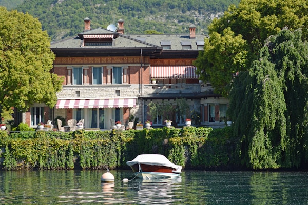 A picturesque lakeside villa surrounded by lush greenery and trees, with a red and white striped awning. The villa has a brick and stone facade, large windows, and outdoor seating. In front, a small boat is moored on calm water, reflecting the serene environment.