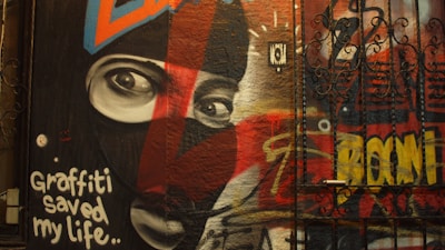 A vibrant piece of graffiti art featuring a large portrait of a person wearing a mask with intense eyes. The artwork includes bold letters and phrases, one of which reads 'Graffiti saved my life..'. The background is filled with various swirls and abstract shapes in multiple colors.