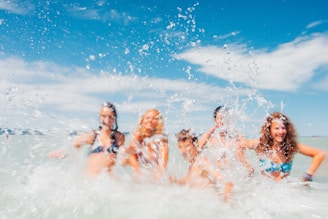 A group of people in swimwear are joyfully splashing in the water, with droplets caught mid-air. The background features a clear blue sky with scattered clouds, contributing to a vibrant and lively scene.
