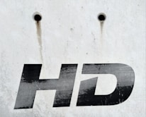 A close-up view of the letters 'HD' printed in bold, black font on a white, weathered surface. Above the text are two small, round holes with rusty streaks trailing down from them.