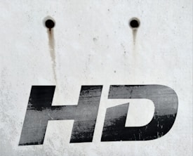 A close-up view of the letters 'HD' printed in bold, black font on a white, weathered surface. Above the text are two small, round holes with rusty streaks trailing down from them.