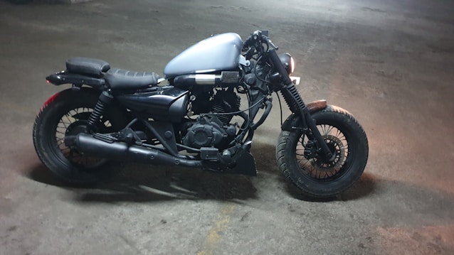 A motorcycle with a minimalist design, featuring a matte black frame, leather seat, and a silver gas tank. The bike is parked on a concrete floor with a dimly lit background, conveying an industrial and rugged aesthetic.