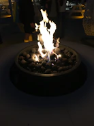 A cozy outdoor setting features a round fire pit with a vibrant and robust flame. Smooth stones are arranged around the flames, enhancing the rustic charm. In the dimly lit background, there are people standing, some picnic tables, and large hanging lanterns.