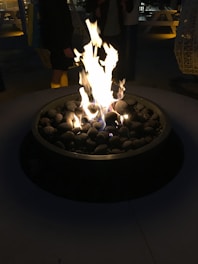 A cozy outdoor setting features a round fire pit with a vibrant and robust flame. Smooth stones are arranged around the flames, enhancing the rustic charm. In the dimly lit background, there are people standing, some picnic tables, and large hanging lanterns.
