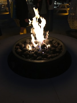 A cozy outdoor setting features a round fire pit with a vibrant and robust flame. Smooth stones are arranged around the flames, enhancing the rustic charm. In the dimly lit background, there are people standing, some picnic tables, and large hanging lanterns.