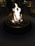 A cozy outdoor setting features a round fire pit with a vibrant and robust flame. Smooth stones are arranged around the flames, enhancing the rustic charm. In the dimly lit background, there are people standing, some picnic tables, and large hanging lanterns.
