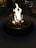 A cozy outdoor setting features a round fire pit with a vibrant and robust flame. Smooth stones are arranged around the flames, enhancing the rustic charm. In the dimly lit background, there are people standing, some picnic tables, and large hanging lanterns.
