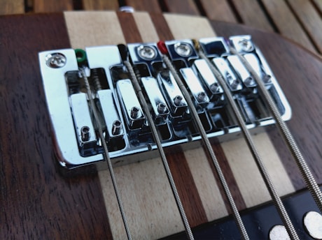 A close-up view of an electric guitar bridge, featuring metallic strings tightly wound over chrome-plated saddles. The wooden surface beneath the bridge showcases a rich combination of brown and beige tones with a striped pattern.