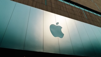 A large, reflective building facade featuring a prominent and shiny apple logo. The facade is composed of smooth, metallic panels with a modern architectural style. The upper portion of the building consists of a more textured, darker material that contrasts with the sleek panels below.