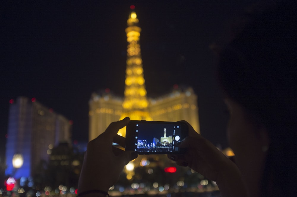 person holding black smartphone taking photo of lighted tower during night time