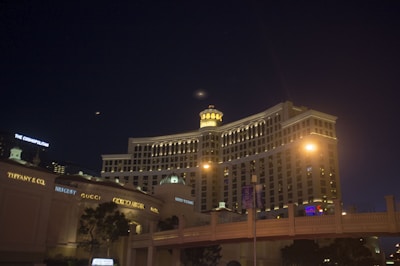 A night view of a large, well-lit hotel with distinctive architecture. Several luxury brand stores are visible on the left side. The sky is dark, with small lights from the surrounding cityscape. Streetlights and building lights illuminate the scene.