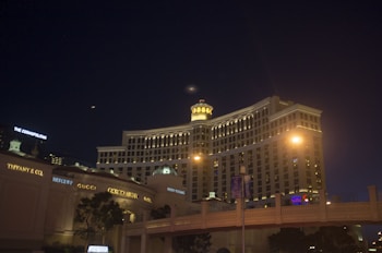 A night view of a large, well-lit hotel with distinctive architecture. Several luxury brand stores are visible on the left side. The sky is dark, with small lights from the surrounding cityscape. Streetlights and building lights illuminate the scene.