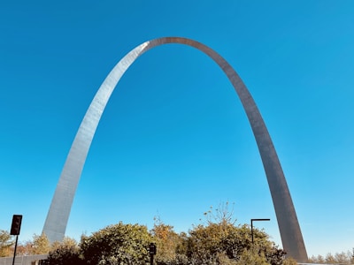 A large stainless steel arch set against a clear blue sky, with greenery and small structures at the base.