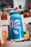 A person is holding a large canister of Lysol Advanced Disinfecting Wipes. The container is prominently blue and white, featuring text that highlights its germ-killing properties and enhanced cleaning power. In the background, there are various bottles and jars that appear to be on a kitchen shelf.