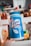 A person is holding a large canister of Lysol Advanced Disinfecting Wipes. The container is prominently blue and white, featuring text that highlights its germ-killing properties and enhanced cleaning power. In the background, there are various bottles and jars that appear to be on a kitchen shelf.