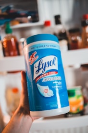 A person is holding a large canister of Lysol Advanced Disinfecting Wipes. The container is prominently blue and white, featuring text that highlights its germ-killing properties and enhanced cleaning power. In the background, there are various bottles and jars that appear to be on a kitchen shelf.