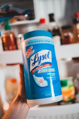A person is holding a large canister of Lysol Advanced Disinfecting Wipes. The container is prominently blue and white, featuring text that highlights its germ-killing properties and enhanced cleaning power. In the background, there are various bottles and jars that appear to be on a kitchen shelf.