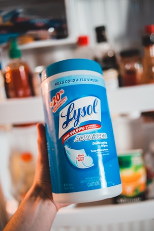 A person is holding a large canister of Lysol Advanced Disinfecting Wipes. The container is prominently blue and white, featuring text that highlights its germ-killing properties and enhanced cleaning power. In the background, there are various bottles and jars that appear to be on a kitchen shelf.