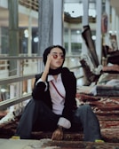 A person wearing a black headscarf, sunglasses, black blazer, and white shirt is sitting on ornamental rugs. The setting appears to be indoors with a modern, industrial feel, possibly in a market or store with rugs stacked in the background. The person has a relaxed and stylish demeanor.