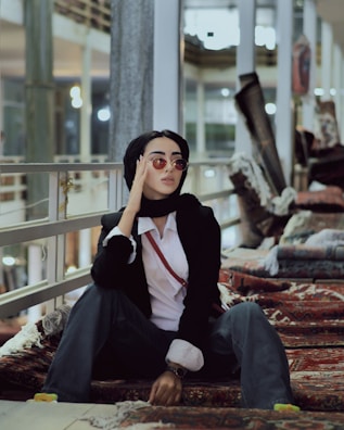 A person wearing a black headscarf, sunglasses, black blazer, and white shirt is sitting on ornamental rugs. The setting appears to be indoors with a modern, industrial feel, possibly in a market or store with rugs stacked in the background. The person has a relaxed and stylish demeanor.