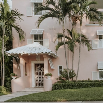 A pastel pink house with a white tiled roof and decorative white shutters is partially surrounded by lush green palm trees. The entrance is accentuated by manicured shrubs and potted plants flanking the door. The lawn is well-kept, and the architecture exudes a tropical, inviting charm.