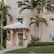 A pastel pink house with a white tiled roof and decorative white shutters is partially surrounded by lush green palm trees. The entrance is accentuated by manicured shrubs and potted plants flanking the door. The lawn is well-kept, and the architecture exudes a tropical, inviting charm.