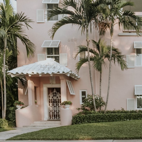 A pastel pink house with a white tiled roof and decorative white shutters is partially surrounded by lush green palm trees. The entrance is accentuated by manicured shrubs and potted plants flanking the door. The lawn is well-kept, and the architecture exudes a tropical, inviting charm.
