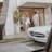 A vintage white Mercedes-Benz parked in front of a stylish boutique. A woman in casual summer attire walks beside a man near the entrance, carrying shopping bags. The scene includes a palm tree and a mannequin visible through the store's glass window.