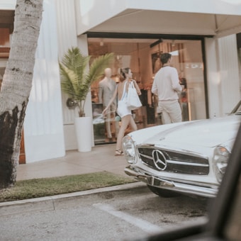A vintage white Mercedes-Benz parked in front of a stylish boutique. A woman in casual summer attire walks beside a man near the entrance, carrying shopping bags. The scene includes a palm tree and a mannequin visible through the store's glass window.