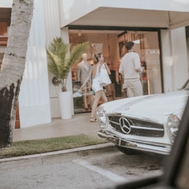 A vintage white Mercedes-Benz parked in front of a stylish boutique. A woman in casual summer attire walks beside a man near the entrance, carrying shopping bags. The scene includes a palm tree and a mannequin visible through the store's glass window.