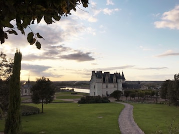 A picturesque landscape featuring a historic castle surrounded by a lush green lawn. A winding cobblestone path leads the eye toward the main building, characterized by its towers and intricate architecture. Trees border the scene, and the sky is a serene mix of clouds and blue, with the setting sun casting a warm glow over the area.