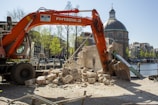 An excavator is actively demolishing a stone structure near a canal. The backdrop includes classic European architecture and a prominent domed building. The setting appears to be urban, with cobblestone paths and a clear blue sky.