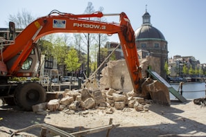 An excavator is actively demolishing a stone structure near a canal. The backdrop includes classic European architecture and a prominent domed building. The setting appears to be urban, with cobblestone paths and a clear blue sky.