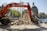 An excavator is actively demolishing a stone structure near a canal. The backdrop includes classic European architecture and a prominent domed building. The setting appears to be urban, with cobblestone paths and a clear blue sky.