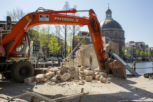 An excavator is actively demolishing a stone structure near a canal. The backdrop includes classic European architecture and a prominent domed building. The setting appears to be urban, with cobblestone paths and a clear blue sky.
