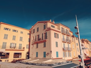 A bright, picturesque street scene in Saint-Tropez featuring colorful, Mediterranean-style buildings with wooden shutters and balconies. The sky is clear and deep blue, enhancing the vibrant atmosphere. Outdoor seating is present, suggesting a brasserie or café setting, with a prominent sign that reads 'LA SARDINE.'