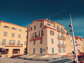 A bright, picturesque street scene in Saint-Tropez featuring colorful, Mediterranean-style buildings with wooden shutters and balconies. The sky is clear and deep blue, enhancing the vibrant atmosphere. Outdoor seating is present, suggesting a brasserie or caf&eacute; setting, with a prominent sign that reads 'LA SARDINE.'