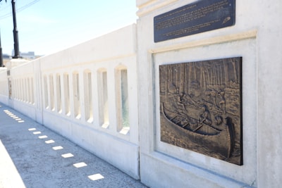 A relief sculpture depicting a historical scene with figures in a canoe, surrounded by a forest and possibly tents or huts in the background. The artwork is mounted on a concrete wall that appears to be part of a bridge or pedestrian walkway. A plaque with text is positioned above the relief.
