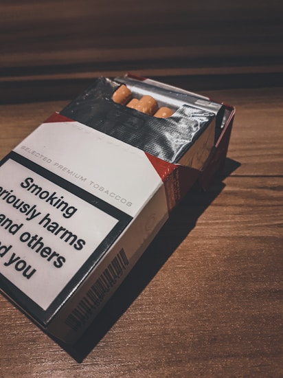 A partially open cigarette pack with a prominent health warning visible on its side. The pack is placed on a wooden surface, and a few cigarettes are partially visible inside the packaging.